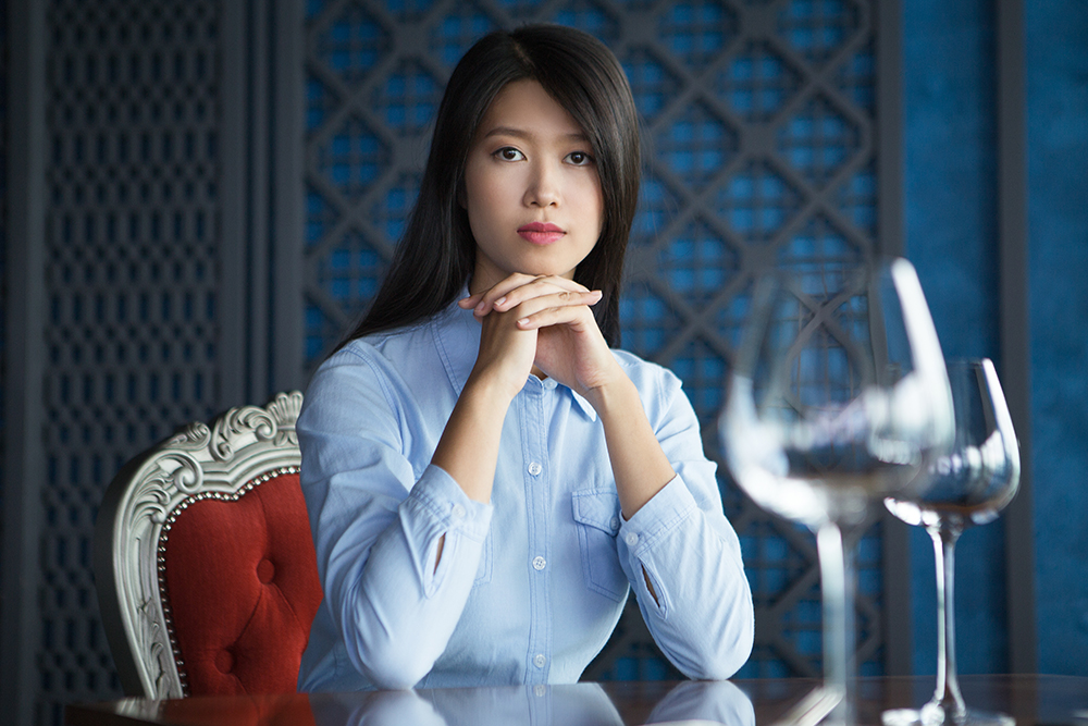Portrait of pretty young Asian woman with her hands under chin sitting at table in luxury restaurant, looking at camera